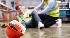 Ein Arbeiter ist hingefallen. Ein anderer kommt diesem zur Hilfe. Im Vordergrund ist ein Schutzhelm zu sehen.