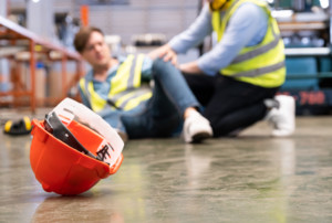 Ein Arbeiter ist hingefallen. Ein anderer kommt diesem zur Hilfe. Im Vordergrund ist ein Schutzhelm zu sehen.