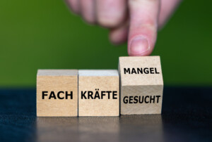 Drei nebeneinanderliegende Holzwürfel. Auf dem ersten Würfel steht "FACH" auf dem zweiten steht "KRÄFTE" und der letzte Würfel wird von einem Finger umgedreht so dass auf der Vorderseite "MANGEL" und auf der Unterseite "GESUCHT" steht.
