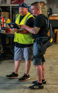 Zwei Männer - einer in Warnweste, der andere mit Exoskelett - gucken auf ein Tablett.