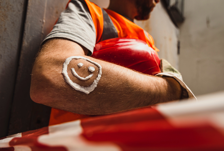 Ein Bauarbeiter lehnt sich an eine Wand und hat ein Smiley aus Sonnencrem auf seinem Arm.