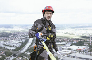 Ein Höhenarbeiter mit Schutzhelm und Sicherheitsgurt steht hoch über einer Stadtlandschaft. Er trägt eine graue Arbeitsjacke mit schwarzen und gelben Details sowie Handschuhe. In seiner Hand hält er eine Sicherungsvorrichtung, die an einem gelben Gurt befestigt ist. Im Hintergrund sind eine Stadt mit Fluss, Straßen und Gebäude aus der Vogelperspektive zu sehen.