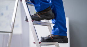 Eine Person in blauer Arbeitshose und schwarzen Arbeitsschuhen steht auf einer Leiter.