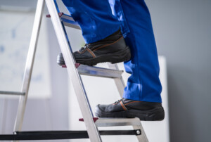 Eine Person in blauer Arbeitshose und schwarzen Arbeitsschuhen steht auf einer Leiter.