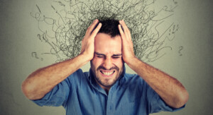 Ein Mann hält sich mit beiden Händen den Kopf und verzieht sein Gesicht, als ob er unter starkem Stress oder Verwirrung leidet. Aus seinem Kopf sprießen wirre, verworrene Linien und Fragezeichen. Der Hintergrund ist grau.