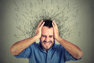 Ein Mann hält sich mit beiden Händen den Kopf und verzieht sein Gesicht, als ob er unter starkem Stress oder Verwirrung leidet. Aus seinem Kopf sprießen wirre, verworrene Linien und Fragezeichen. Der Hintergrund ist grau.