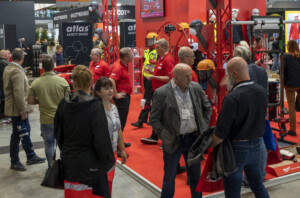 Messebesucher auf einer Veranstaltung stehen in einem Stand von Atlas Safety Shoes, der Sicherheitskleidung und -ausrüstung wie Helme und Schutzkleidung zeigt. Die meisten Personen tragen rote T-Shirts und Namensschilder. Im Hintergrund sind weitere Besucher und Aussteller zu sehen.