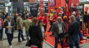 Messebesucher auf einer Veranstaltung stehen in einem Stand von Atlas Safety Shoes, der Sicherheitskleidung und -ausrüstung wie Helme und Schutzkleidung zeigt. Die meisten Personen tragen rote T-Shirts und Namensschilder. Im Hintergrund sind weitere Besucher und Aussteller zu sehen.
