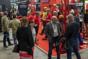 Messebesucher auf einer Veranstaltung stehen in einem Stand von Atlas Safety Shoes, der Sicherheitskleidung und -ausrüstung wie Helme und Schutzkleidung zeigt. Die meisten Personen tragen rote T-Shirts und Namensschilder. Im Hintergrund sind weitere Besucher und Aussteller zu sehen.
