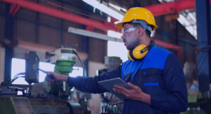 Ein Arbeiter mit gelbe Schutzhelm und Ohrenschützern um seinen Nacken, hält ein Tablet in seiner Hand und guckt auf die Anzeige einer Arbeitsmaschine.