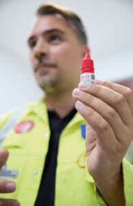 Ein Mann in gelber Arbeitsjacke hält ein Fläschschen mit rotem Deckel hoch. Darauf sind Gefahrensymbole zu erkennen.