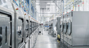 Blick in eine industrielle Wäscherei mit mehreren Waschmaschinen aus Edelstahl, die in Reihen aufgestellt sind. Zwischen den Maschinen sind weitere Geräte und Wäschewagen zu sehen.