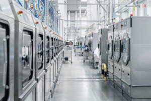 Blick in eine industrielle Wäscherei mit mehreren Waschmaschinen aus Edelstahl, die in Reihen aufgestellt sind. Zwischen den Maschinen sind weitere Geräte und Wäschewagen zu sehen.