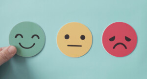 Drei runde Emoticons auf einem blauen Hintergrund: Ein grünes, lachendes Gesicht, ein gelbes, neutral aussehendes Gesicht und ein rotes, trauriges Gesicht. Eine Hand legt das grüne Emoticon gerade ab.