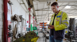 Ein Mann mit gelber Arbeitsjacke steht in einer Werkstatt und guckt auf sein Handy.