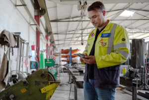 Ein Mann mit gelber Arbeitsjacke steht in einer Werkstatt und guckt auf sein Handy.