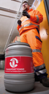 Eine graue Transporttonne mit rotem Warnaufkleber steht im Vordergrund. Der Aufkleber trägt die Aufschrift „TRANSPORTTONNE FÜR DEFEKTE HOCHENERGIEBATTERIEN“ sowie ein Symbol für Batterierecycling. Im Hintergrund steht eine Person in orangefarbener Warnschutzkleidung mit reflektierenden Streifen. Sie trägt Handschuhe und scheint eine Erklärung zu geben. Die Umgebung wirkt wie ein Lagerraum mit Regalen und Kartons.