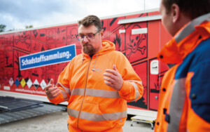 Zwei Männer in Warnschutzkleidung stehen vor einem Container mit der Aufschrift "Schadstofsammlung" und unterhalten sich.
