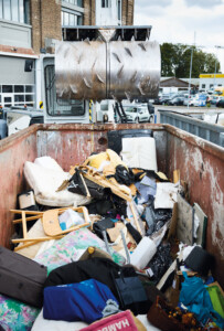 Ein großer Container ist mit Sperrmüll gefüllt, darunter Matratzen, Möbelstücke, Taschen und verschiedene Abfälle. Darüber schwebt eine massive, metallene Walze mit Zacken, die offenbar zur Verdichtung des Mülls dient. Im Hintergrund sind eine Lagerhalle, Autos und Müllcontainer zu sehen.