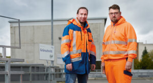 Zwei Männer in Arbeitskleidung stehen auf einem Betriebsgelände. Der Mann links trägt eine blau-orange Warnschutzjacke und Jeans, während der Mann rechts eine vollständig orangefarbene Warnschutzkleidung mit reflektierenden Streifen trägt. Beide blicken in die Kamera. Im Hintergrund sind ein Gebäude mit der Aufschrift „Grünschnitt“ sowie ein Metallgeländer zu sehen. Der Himmel ist bewölkt.