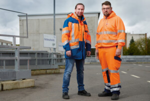 Zwei Männer in Arbeitskleidung stehen auf einem Betriebsgelände. Der Mann links trägt eine blau-orange Warnschutzjacke und Jeans, während der Mann rechts eine vollständig orangefarbene Warnschutzkleidung mit reflektierenden Streifen trägt. Beide blicken in die Kamera. Im Hintergrund sind ein Gebäude mit der Aufschrift „Grünschnitt“ sowie ein Metallgeländer zu sehen. Der Himmel ist bewölkt.