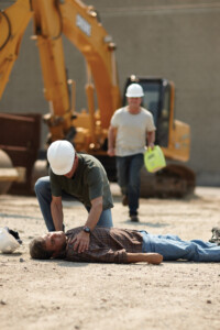 Ein Mann liegt bewusstlos auf dem Boden. Ein anderer Mann mit weißem Bauhelm, kniet über ihn. Im Hintergrund ist ein gelber Bagger zu sehen und ein weiterer Mann mit weißem Bauhelm und einem Defibrillator in der linken Hand rennt in Richtung der beiden anderen Männer.