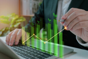 Ein Mann in Anzug arbeitet an einem Laptop, während er mit einem Stift eine virtuelle, grüne Wachstumsstatistik verfolgt. Die Grafik zeigt aufsteigende Balken mit kleinen Blättern an der Spitze, die Nachhaltigkeit und wirtschaftliches Wachstum symbolisieren. Im Hintergrund sind Pflanzen und ein Büroambiente zu erkennen.