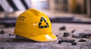 Ein gelber Bauhelm mit einem Recycling Logo liegt auf einem asphaltierten Boden. Neben dem Bauhelm liegen kleine Steine und Stücke des Asphalts.