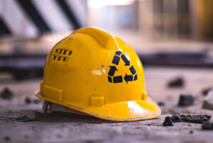 Ein gelber Bauhelm mit einem Recycling Logo liegt auf einem asphaltierten Boden. Neben dem Bauhelm liegen kleine Steine und Stücke des Asphalts.