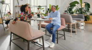 Moderner Arbeitsplatz mit großen Fenstern und viel Tageslicht. Zwei Personen, eine Frau mit lockigem Haar und bunter Bluse sowie ein Mann mit grauem Haar und hellblauem Hemd, sitzen an einem Tisch und unterhalten sich. Im Hintergrund arbeiten weitere Menschen an Laptops, eine Person bestellt an der Theke. Die Einrichtung ist stilvoll mit gepolsterten Sitzbänken, Pflanzen und hellen Farben
