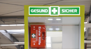 Bildausschnitt einer Leuchtend grüne Erste-Hilfe-Station mit der Aufschrift „GESUND + SICHER“ in einer industriellen Umgebung. Die Station enthält einen Erste-Hilfe-Kasten, Notfallinformationen und Aufbewahrungsfächer. Im Hintergrund sind Regale, Maschinen und Werkbänke zu sehen