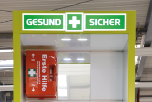 Bildausschnitt einer Leuchtend grüne Erste-Hilfe-Station mit der Aufschrift „GESUND + SICHER“ in einer industriellen Umgebung. Die Station enthält einen Erste-Hilfe-Kasten, Notfallinformationen und Aufbewahrungsfächer. Im Hintergrund sind Regale, Maschinen und Werkbänke zu sehen