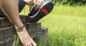 Ein barfüßiger Mann sitzt auf einem Haufen Pflastersteine und zieht sich einen Barfußschuh am linken Fuß an. Sein rechter Fuß, mit drei Lederfußbändern geschmückt, stützt sich an den Steinen ab. Im Hintergrund sind Rasen und Büsche zu sehen.