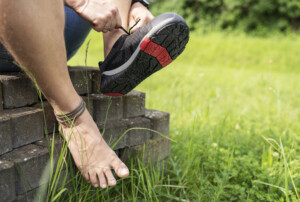Ein barfüßiger Mann sitzt auf einem Haufen Pflastersteine und zieht sich einen Barfußschuh am linken Fuß an. Sein rechter Fuß, mit drei Lederfußbändern geschmückt, stützt sich an den Steinen ab. Im Hintergrund sind Rasen und Büsche zu sehen.