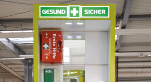 Bildausschnitt einer Leuchtend grüne Erste-Hilfe-Station mit der Aufschrift „GESUND + SICHER“ in einer industriellen Umgebung. Die Station enthält einen Erste-Hilfe-Kasten, Notfallinformationen und Aufbewahrungsfächer. Im Hintergrund sind Regale, Maschinen und Werkbänke zu sehen