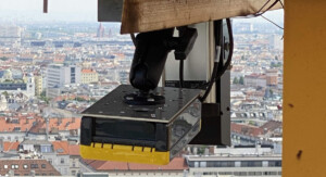 Eine Sensorik-Einheit ist an der Struktur eines Baukrans montiert und ragt nach unten. Im Hintergrund erstreckt sich eine Stadtlandschaft mit Gebäuden und roten Dächern.