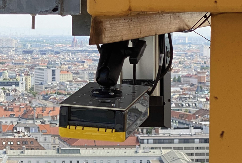 Eine Sensorik-Einheit ist an der Struktur eines Baukrans montiert und ragt nach unten. Im Hintergrund erstreckt sich eine Stadtlandschaft mit Gebäuden und roten Dächern.