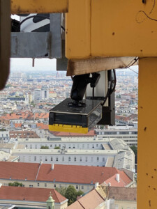 Eine Sensorik-Einheit ist an der Struktur eines Baukrans montiert und ragt nach unten. Im Hintergrund erstreckt sich eine Stadtlandschaft mit Gebäuden und roten Dächern.