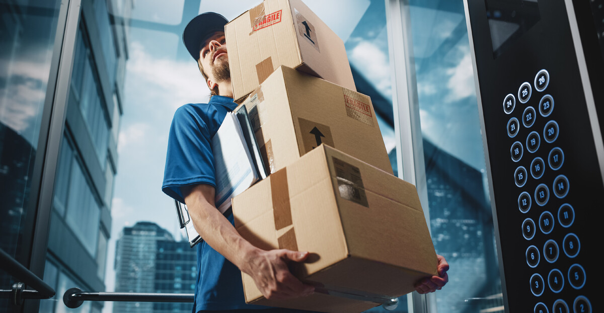 Ein Paketbote mit blauer Uniform, steht in einem verglasten Fahrstuhl und hält drei Pakete und ein Klemmbrett unter seinem Arm. Im Hintergrund sieht man einige Wolkenkratzer.