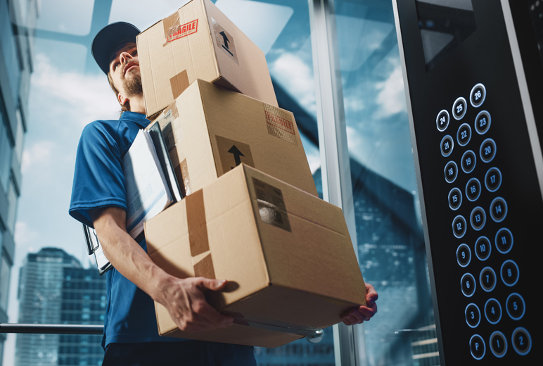 Ein Paketbote mit blauer Uniform, steht in einem verglasten Fahrstuhl und hält drei Pakete und ein Klemmbrett unter seinem Arm. Im Hintergrund sieht man einige Wolkenkratzer.