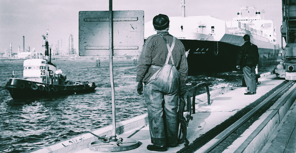 Schwarz-weißes Bild eines Hafens. Im Mittelpunkt steht ein Mann, der von hinten zu sehen ist, neben einem Schild und hält ein Seil in der Hand. Im Hintergrund ist ein weiterer Mann zu sehen. Auf dem Wasser schwimmen ein großes und ein kleines Schiff.