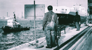 Schwarz-weißes Bild eines Hafens. Im Mittelpunkt steht ein Mann, der von hinten zu sehen ist, neben einem Schild und hält ein Seil in der Hand. Im Hintergrund ist ein weiterer Mann zu sehen. Auf dem Wasser schwimmen ein großes und ein kleines Schiff.