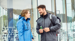 Ein Mann in schwarzer Softshell Jacke und eine Frau in blauer Softshell Jacke stehen draußen vor einer Fensterfront und unterhalten sich. Der Mann trägt einen grauen Rucksack und hält eine grüne Getränkedose.