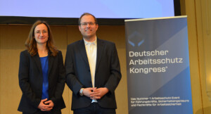 Ein Mann und eine Frau posieren vor einem Aufsteller mit der Aufschrift "Deutscher Arbeitsschutz Kongress".