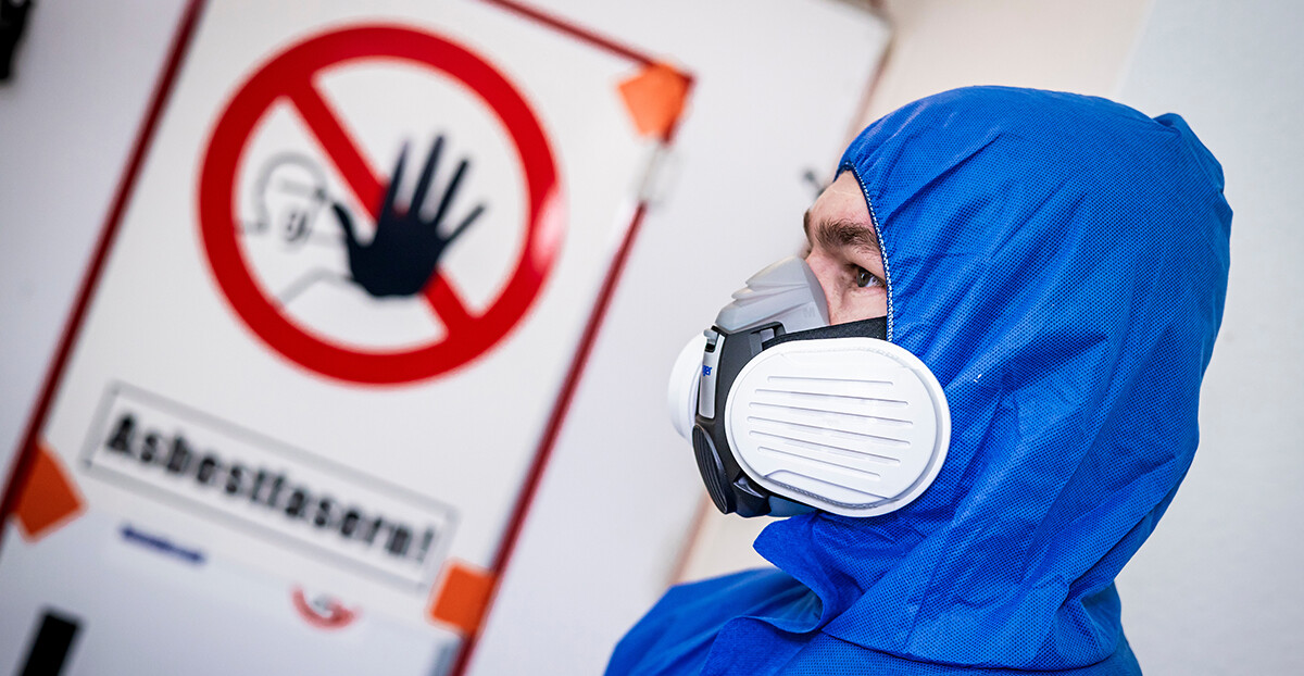 Eine Person im blauen Einwegoverall trägt eine Halbmaske mit Partikelfiltern und steht vor einer Tür mit durchgestrichenem Handsymbol und dem Warnhinweis „Asbestfasern!“.
