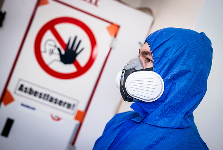 Eine Person im blauen Einwegoverall trägt eine Halbmaske mit Partikelfiltern und steht vor einer Tür mit durchgestrichenem Handsymbol und dem Warnhinweis „Asbestfasern!“.