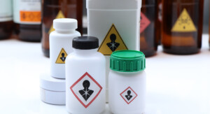 Mehrere weiße Kunststoffbehälter und braune Glasflaschen stehen dicht beieinander auf einer weißen Fläche, alle mit dem GHS-Piktogramm „Gesundheitsgefahr“ gekennzeichnet.