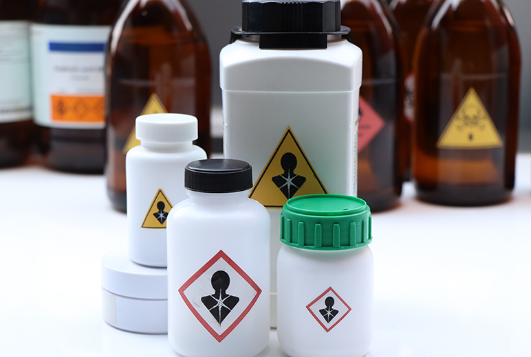 Mehrere weiße Kunststoffbehälter und braune Glasflaschen stehen dicht beieinander auf einer weißen Fläche, alle mit dem GHS-Piktogramm „Gesundheitsgefahr“ gekennzeichnet.