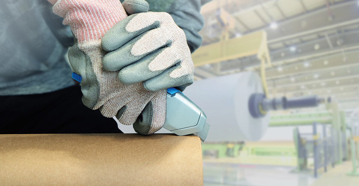 Ein Arbeiter mit schnittfesten Handschuhen setzt ein Sicherheitsmesser an die Papierhülse einer großen Rolle in einer hell ausgeleuchteten Produktionshalle.