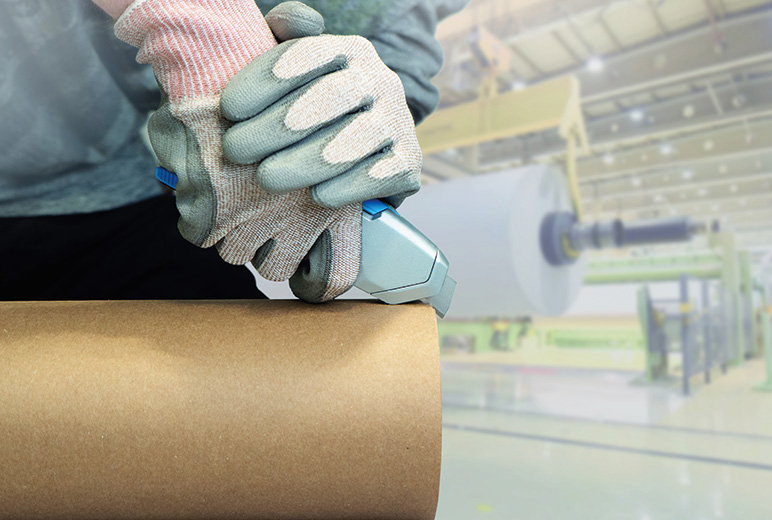 Ein Arbeiter mit schnittfesten Handschuhen setzt ein Sicherheitsmesser an die Papierhülse einer großen Rolle in einer hell ausgeleuchteten Produktionshalle.